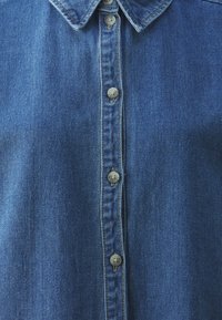 Blå denimskjorte med spiss krage, knappestolpe foran med sølvknapper og synlige sømmer, som viser et strukturert stoff.