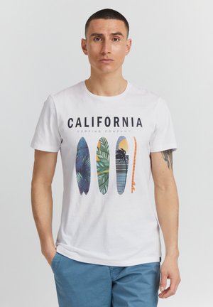 Man with short dark hair wearing white T-shirt with "CALIFORNIA" and four colorful surfboard graphics, paired with blue shorts against plain background.