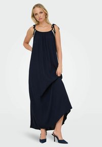 Robe maxi bleu marine avec un design fluide, des bretelles à nouer ajustables et un tissu texturé. Assortie à des talons hauts bleu foncé.