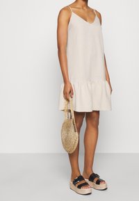 Kvinna i en beige ärmlös klänning som håller en rund vävd halmhandväska, parat med svarta plattformssandaler med spännen.