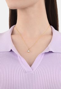 Collar de cadena delicada con colgante de oro que presenta una piedra clara en forma de lágrima, llevada sobre una camiseta de cuello de canalé de color púrpura claro.