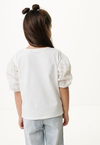 Camiseta blanca de algodón con mangas abullonadas, hecha de tela bordada. La vista posterior muestra un escote redondo simple y jeans de mezclilla azul claro.