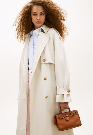 Vrouw met krullend haar, gekleed in een beige trenchcoat over een blauw shirt en een bruine leren handtas met gouden accenten vasthoudend.