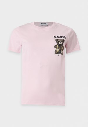 Kobieta stoi z ręką w kieszeni, ubrana w różowy t-shirt, czarną mini spódnicę z różowym futrzanym wykończeniem i czarne sandały na obcasie.
