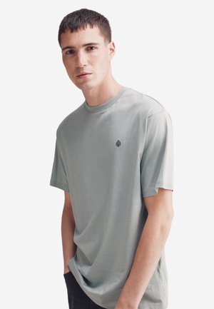 Hellgrünes Baumwoll-T-Shirt mit Rundhalsausschnitt und kurzen Ärmeln. Besitzt ein kleines Baum-Logo auf der linken Brustseite. Einfarbig, glatte Textur.