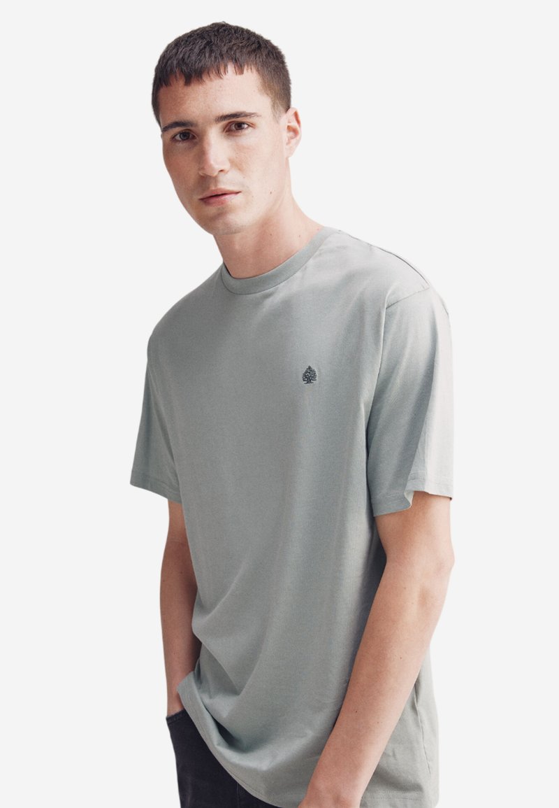 T-shirt in cotone verde chiaro con scollo rotondo e maniche corte. Presenta un piccolo logo ad albero sull'area sinistra del petto. Colore uniforme, texture liscia.