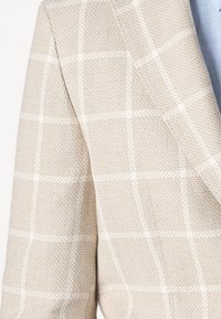 Beżowa marynarka w kratę z teksturowanej tkaniny, z białymi liniami w wzór siatki i szerokim wzorem klap.