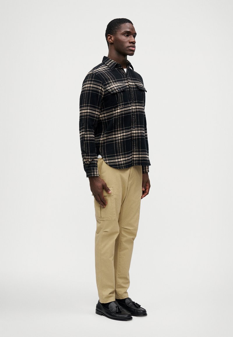 Camisa de franela a cuadros en negro y marrón con dos bolsillos en el pecho, pantalones cargo color caqui y mocasines negros. El modelo se encuentra de lado contra un fondo neutro.