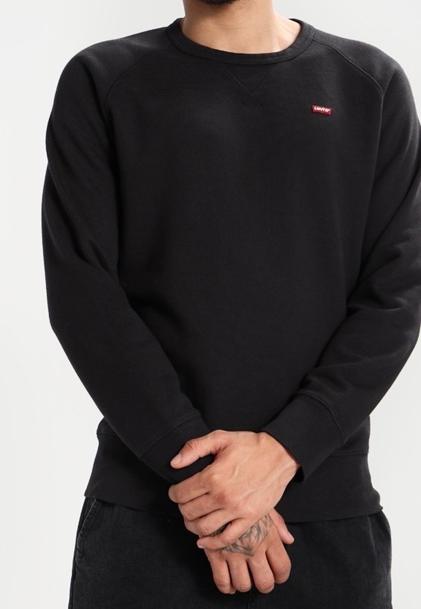 Homme portant un sweat Levi’s noir uni, mains jointes devant lui, montrant un tatouage sur la main gauche.