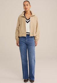 Beige bomber bunda s vysokým límcem, kombinovaná s bílým svetr s výstřihem do V a námořnickými pruhy, a světle modrými širokými kalhotami.
