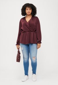 A young woman stands confidently, wearing a burgundy wrap blouse, distressed jeans, and white sneakers, holding a small handbag.