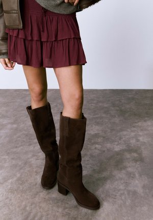 Botas de ante marrón hasta la rodilla con un tacón de bloque, que presentan una textura suave y un sutil detalle de costura, combinadas con una falda burgundy en capas.