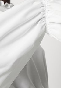 Blouse blanche avec poignet froncé et tissu léger et lisse. Présente une coupe décontractée et un léger éclat, mettant en valeur la fluidité et la chute du tissu.