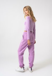 Cropped top viola a maniche lunghe con design aderente, abbinato a pantaloni della tuta viola con vita elastica e tasche laterali. Sneakers bianche.