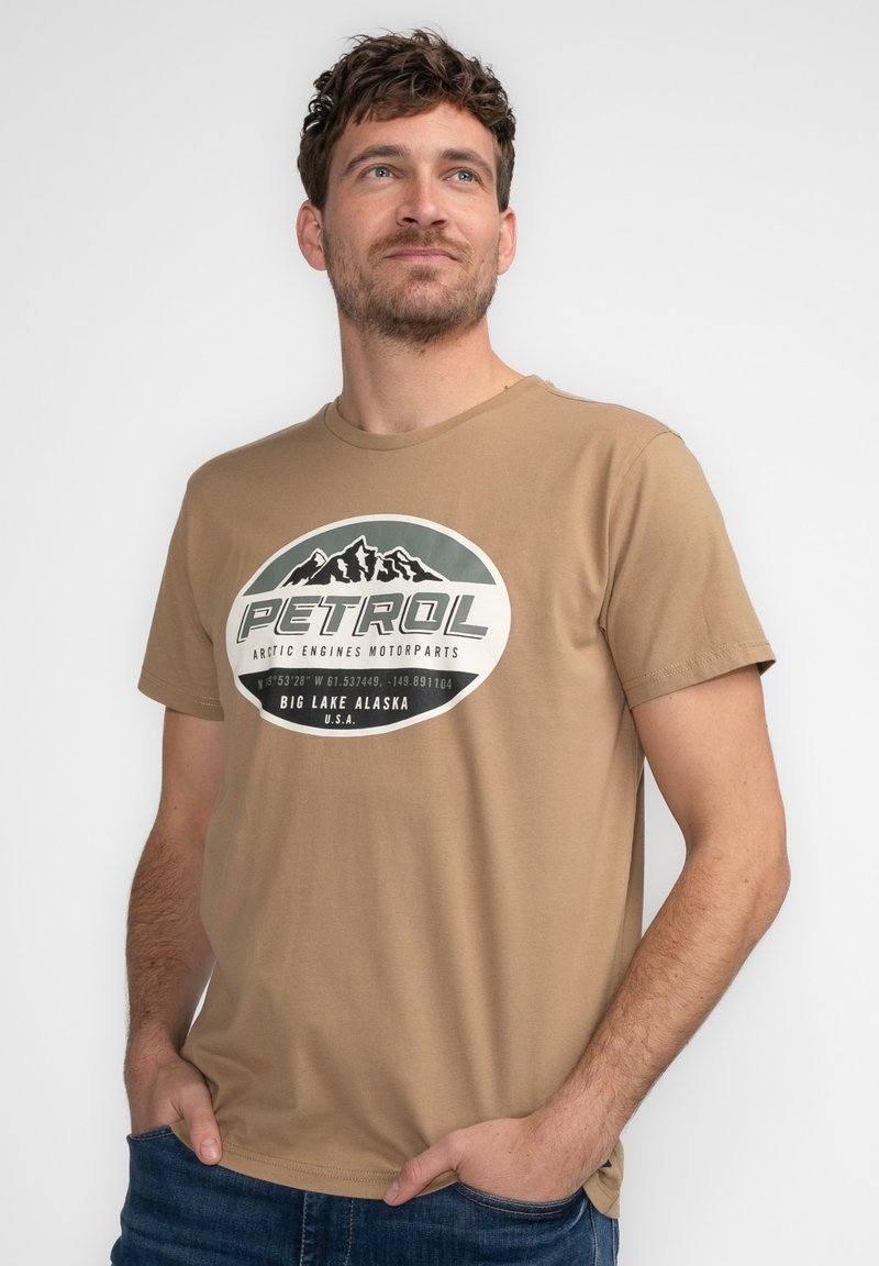 Brun bomulds T-shirt med et rundt trykt logo med bjerge og ordet "PETROL", parret med blå jeans. Korte ærmer og rund hals.