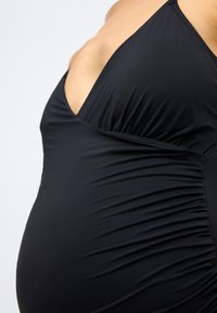 Prim-plan al unui burtă de gravidă îmbrăcată într-un costum de baie matern negru, cu pliuri și decolteu adânc, pe un fundal simplu.