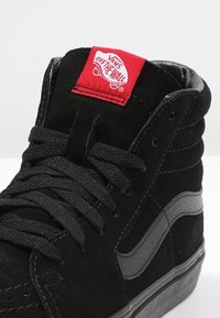 Černé vysoké tenisky z semiše s koženým detailem. Mají ploché černé šňůrky a červenou visačku s logem Vans Off The Wall.