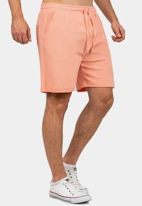 Pantaloni corti color pesco chiaro realizzati in tessuto morbido, con vita regolabile mediante cordoncino, tasche laterali e una vestibilità comoda. Abbinati a sneakers bianche.