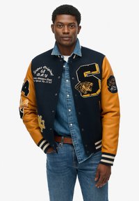 Chaqueta universitaria de la marina con mangas de cuero naranja, que presenta parches bordados y rayas en los puños. Llevada sobre una camisa de mezclilla y jeans.