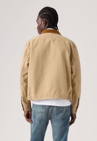 Χακί βαμβακερό μπουφάν με καφέ γιακά από κορντουρ, άνετη εφαρμογή, χαμηλωμένους ώμους και ριμπ μανσέτες. Φοριέται πάνω από λευκό πουκάμισο και μπλε τζιν.