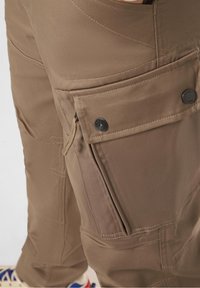 Hnědé cargo kalhoty s více kapsami, zipovou kapsou a zesílenými švy. Vyrobené z odolné látky s hladkou texturou.