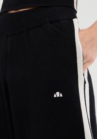 Schwarze Jogginghosen mit weißen Seitenstreifen und einem kleinen weißen Logo am linken Oberschenkel. Das Material wirkt weich mit einer strukturierten Oberfläche.