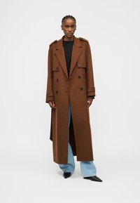 Γυναίκα που φοράει μακρύ καφέ παλτό διπλής στερέωσης, μαύρο τοπ, ανοιχτό γαλάζιο τζιν με φαρδιά μπατζάκια και μαύρα μυτερά παπούτσια, στέκεται μπροστά σε απλό φόντο.