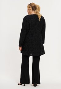 Sort, blomstermønstret lang frakke med lange ærmer og asymmetrisk kant. Kombination med lige sorte bukser og åbne sko.