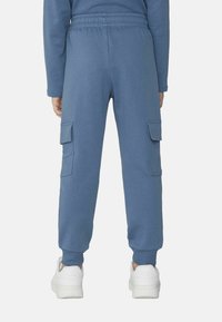 Modré tepláky z bavlněné směsi, které mají dvě boční cargo kapsy, elastický pas a manžetové zakončení. Nosič má na sobě bílé tenisky.