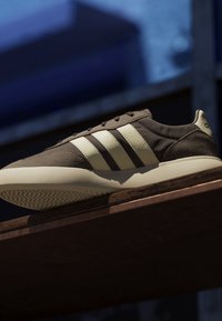 Bruine en beige sneaker met drie witte strepen, beige zool, rustend op een houten oppervlak tegen een wazige donkerblauwe achtergrond.