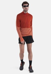 Maglia a maniche lunghe con zip in gradiente arancione, caratterizzata da un tessuto testurizzato e aperture per i pollici, abbinata a pantaloni corti neri e scarpe da ginnastica.
