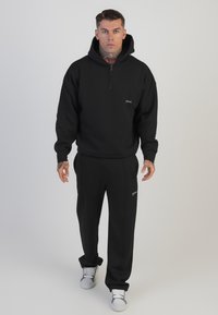Svart zip-up hoodie med en framficka, parat med matchande slim-fit joggers. Båda har minimalistisk branding och en mjuk textur.