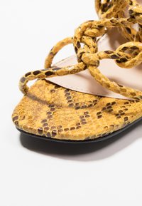 Żółte sandały z wężowej skóry z plecionymi paskami, z gładką cholewką i otwartym noskiem. Czarna gumowa podeszwa z teksturowanym detalem.