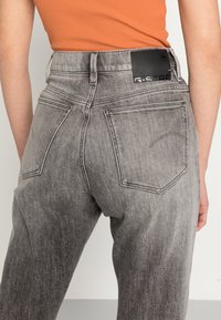Jeans de denim cinza de cintura alta com bolsos traseiros e um patch de couro na cintura. Textura sutil desbotada e corte reto.
