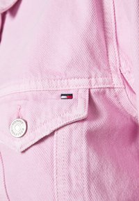 Chaqueta de mezclilla rosa con un botón plateado con la inscripción "TOMMY HILFIGER" grabada. Incluye un pequeño detalle del logo en el bolsillo.