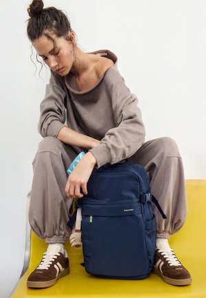 Ženska v bež trenirki in rjavih supergah sedi na rumenem klopci, drži modri nahrbtnik in majhen moder predmet v roki.