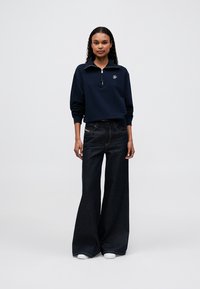 Hanorac bleumarin cu fermoar și guler, asociat cu blugi largi din denim închisi. Outfit-ul are accente minime și adidași albi.