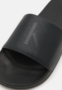 Sandale noire à glissière avec une sangle texturée emboutie du logo Calvin Klein, semelle lisse et design minimaliste.