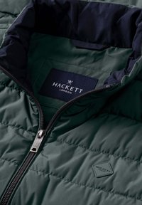 Gewatteerd groen jack met een marineblauwe voering, voorzien van een zilveren ritssluiting en een zwart label met de tekst "HACKETT LONDON" en een logo.