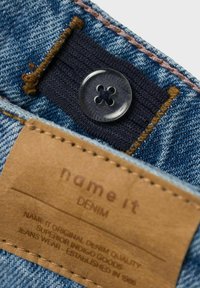 Denim tkanina s plavim pranjem, s crnim gumbom i smešnim kožnim oznakom urezanim s detaljima o brendu i proizvodu.