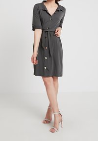 Šedé zavinovací šaty s límcem, krátkými rukávy a opaskem v pase. Mají velké knoflíky na přední straně a hladký, lehký materiál.