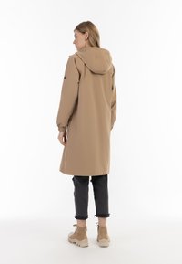 Beige plášť s kapucí a rovný střih, elastické manžety a boční kapsy, spolu s tmavými, roztrhanými džíny a béžovými kotníkovými botami.