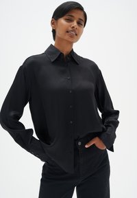 Schwarzes Satin-Hemd mit langen Ärmeln und klassischem Kragen, kombiniert mit dunklen Jeans. Hat eine glatte, glänzende Textur und geknöpfte Manschetten.