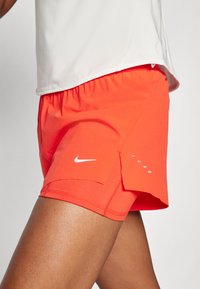 Pantalones cortos de running para mujer de Nike, naranja brillante, tejido ligero, cinturilla elástica, aberturas laterales, detalle del logotipo en blanco, textura suave.