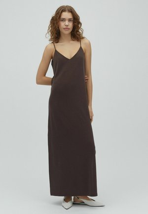 Junge Frau mit lockigem Haar, die ein langes, dunkelbraunes ärmelloses Kleid und weiße spitze flache Schuhe trägt, steht vor einem schlichten hellen Hintergrund.