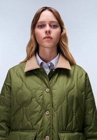 Giacca trapuntata verde oliva con colletto beige, dotata di chiusure a bottoni e tasche. Il motivo testurizzato crea un effetto ondulato.
