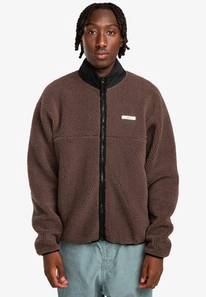 Brown fleece jacket with a black collar, front zip closure, and a rectangular label on the chest. Soft texture and looser fit.