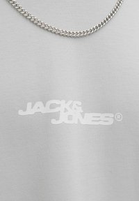 Pelēka auduma t-krekls ar baltu logo "JACK & JONES". Metāla ķēdes kaklarota ir novietota pār dekoltē.