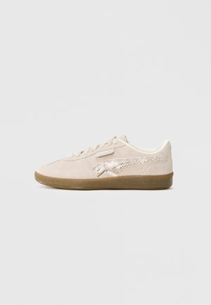 Niski beżowy zamszowy sneaker z wiązaniem na sznurówki, teksturowanym metalicznym paskiem z boku oraz brązową gumową podeszwą na jednolitym tle.