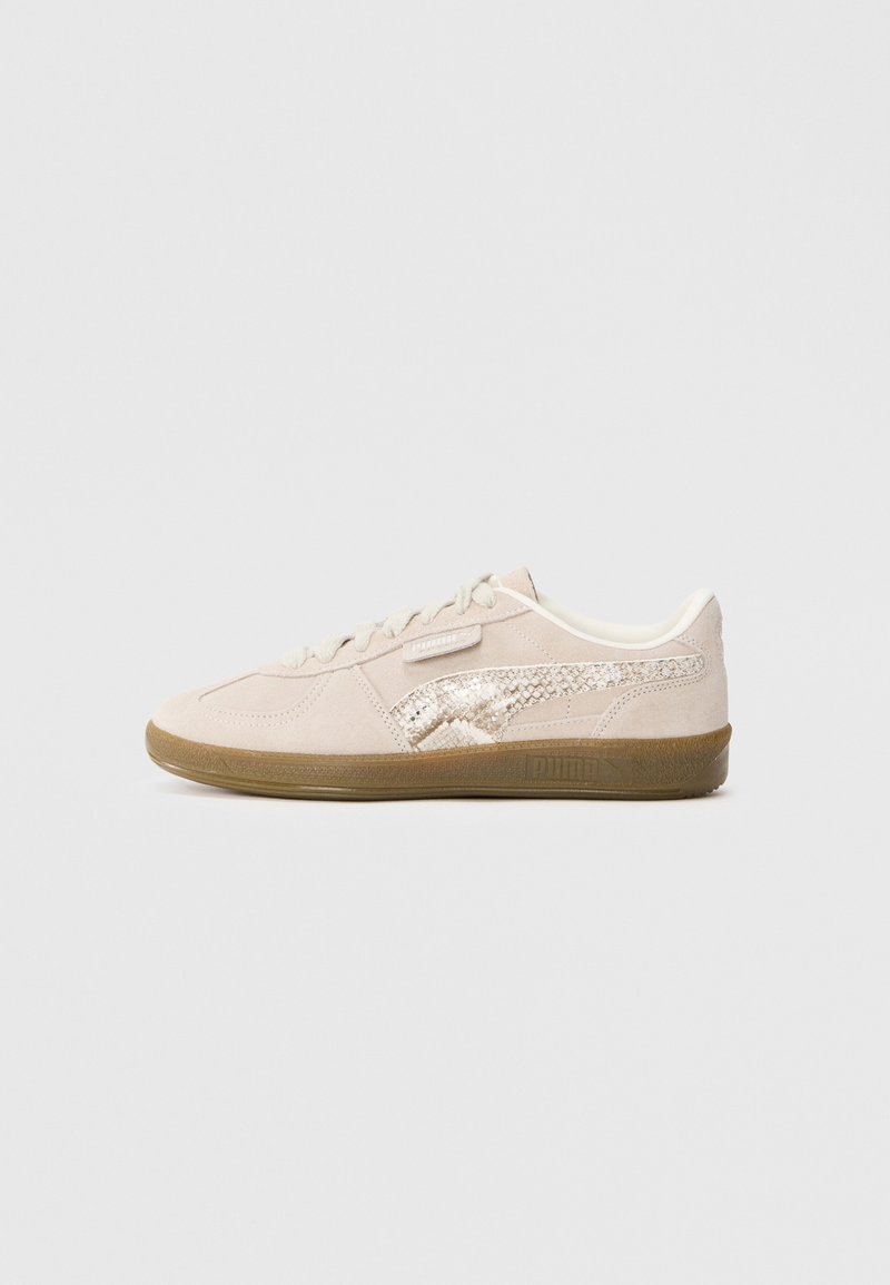 Baskets basses en daim beige avec un design à lacets, bande latérale métallique texturée et semelle en caoutchouc marron sur fond uni.